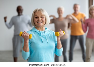 Multiracial group of senior people in sportswear doing strength building fitness exercises with dumbbells, holding fitness tools and smiling at camera, selective focus on positive elderly lady