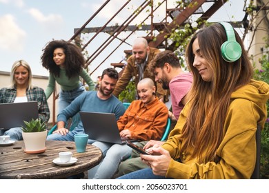 Multiracial group of students working together using digital devices such as laptops and smartphones in the campus terrace, co-working outdoors together. Main focus on the woman with the headphones - Powered by Shutterstock