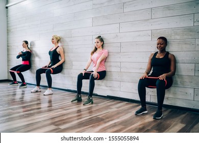 Multiracial Group Of Young Athletic Slim Women Doing Static Exercise With Rubber Bands Leaning On Wall Of Gymnasium In Squat