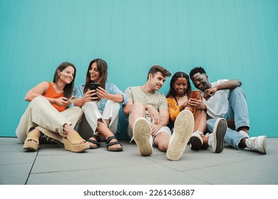 Multiracial group of young friends enjoying and smiling using their mobile phone app sitting at teal wall. Diverse teenagers having fun sharing messages with each other on cellphone at blue clor - Shutterstock ID 2261436887