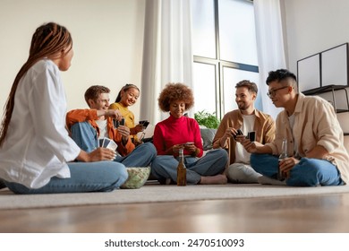 multiracial group of young friends playing cards drinking beer and having fun at home, students sitting on the floor and gambling and drinking alcohol together in the room - Powered by Shutterstock