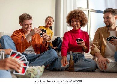 multiracial group of young people sitting at home with beer and popcorn and playing cards with friends, students of different ethnicities playing gambling board games at a party and having fun - Powered by Shutterstock