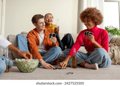 multiracial group of young people sitting at home with beer and popcorn and playing cards with friends, students of different ethnicities playing gambling board games at a party and having fun - Powered by Shutterstock