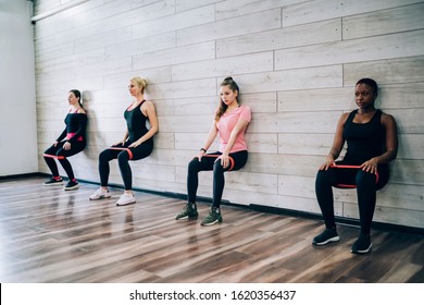 Multiracial Group Of Young Slim Sporty Women Doing Static Exercise With Fitness Rubbers Leaning On Gymnasium Wall In Squat Putting Hands On Knees