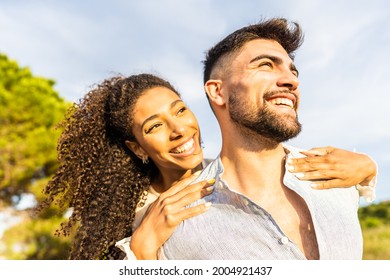 Multiracial Happy Young Beautiful Couple In Love Having Fun Outdoor In Nature At Dusk Looking At Horizon Kissed By The Setting Sun. Toothy Smiling Afro-American Girl Embracing From Back Her Boyfriend