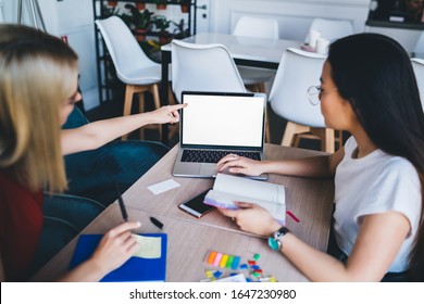 Multiracial ladies  working with laptop sitting at table loaded with notepads markers stickers in modern creative workplace with white chairs opposite of plants in pots on shelving 