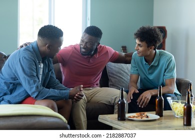 Multiracial Male Friends With Beer Bottles And Food On Table Chatting While Relaxing On Sofa At Home. Alcohol, Unaltered, Friendship, Togetherness, Social Gathering, Enjoyment And Weekend Activities.