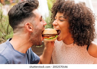 Multiracial man and woman eating tasty hamburger together during romantic date in restaurant in summer