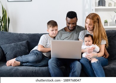 A multiracial parents and their two childs on the sofa with laptop at home, watching movies, web browsing, playing game. A multi ethnic family spend time with with gadget