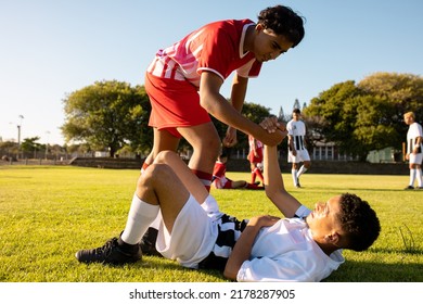 Multiracial player giving hand to injured opponent lying on field in getting up against clear sky. Copy space, playground, match, assistance, pain, unaltered, rivalry, soccer, competition and sport.