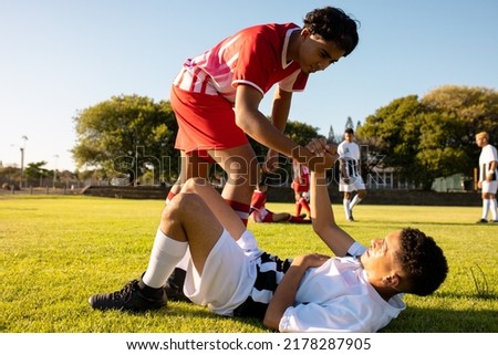 Multiracial player giving hand to injured opponent lying on field in getting up against clear sky. Copy space, playground, match, assistance, pain, unaltered, rivalry, soccer, competition and sport.