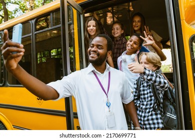 Multiracial pupils and driver smiling and taking selfie photo on cellphone by school bus outdoors