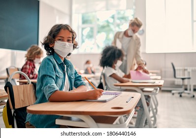 Mehrrassige Schüler der Grundschule sind bereit zu studieren, nach Covid-19 Quarantäne und Schließung. Kinder im Klassenzimmer mit Lehrer, die Gesichtsmasken tragen und Antiseptika zur Koronavirus-Prävention verwenden.