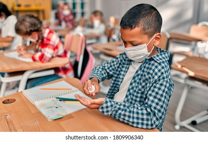 Multiracial pupils of primary school are ready to study after Covid-19 quarantine and lockdown. Children in class room wearing face masks and using antiseptic for coronavirus prevention.