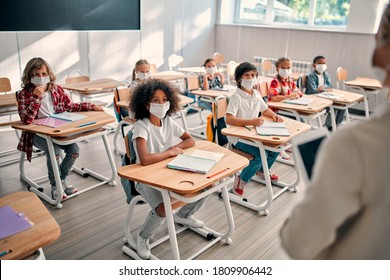 Multiracial pupils of primary school are ready to study after Covid-19 quarantine and lockdown. Children in class room with teacher wearing face masks and using antiseptic for coronavirus prevention.