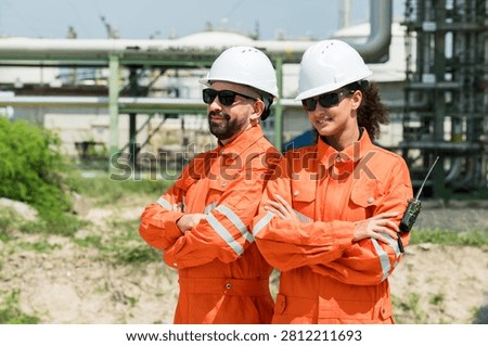Multiracial refinery engineer duo standing with crossed arms near pipeline corridor, carrying walkie talkie during petrochemical safety patrol, presenting confident maintenance workforce readiness