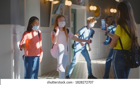 Multiracial Schoolchildren Wearing Facial Mask Relaxing In School Corridor During Break. Diverse Girls Students Filming Dance On Smartphone While Boys Classmates Having Fun On Background