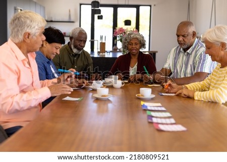 Multiracial senior friends playing bingo while having coffee and cookies at dining table. Nursing home, luck, leisure game, snacks, unaltered, togetherness, support, assisted living, and retirement.