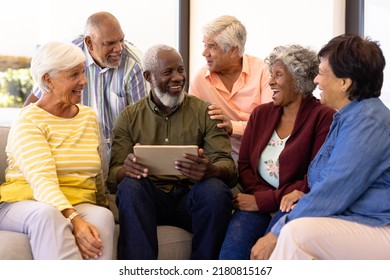 Multiracial senior man holding digital tablet looking at laughing friends while sitting on sofa. Nursing home, wireless technology, unaltered, togetherness, support, assisted living, retirement. - Powered by Shutterstock