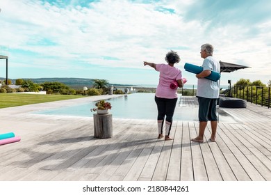 Multiracial Senior Woman Pointing While Standing With Male Friend On Hardwood Floor At Yard. Nursing Home, Swimming Pool, Unaltered, Togetherness, Support, Assisted Living, Retirement, Fitness.