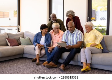 Multiracial Seniors Looking At Digital Tablet Using By Male Friend While Sitting On Sofa. Nursing Home, Wireless Technology, Unaltered, Togetherness, Support, Assisted Living, Retirement Concept.