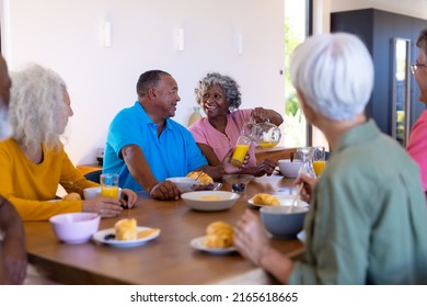 Multiracial Seniors Looking At Happy Woman Serving Juice To Man At Dining Table In Retirement Home. Breakfast, Unaltered, Food, Drink, Healthy, Togetherness, Support, Assisted Living, Retirement.