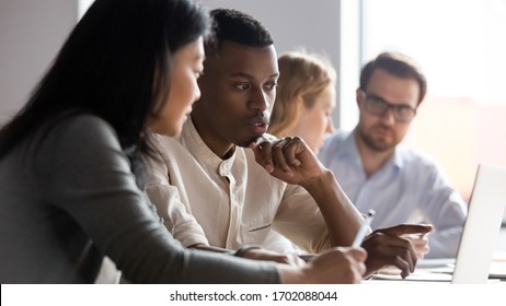 Multiracial serious colleagues sit at desk talk brainstorm using computer cooperate at office briefing, serious diverse coworkers speak discuss business ideas at boardroom meeting together