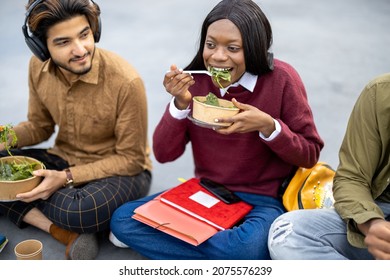 Multiracial Students Sitting And Eating Salad During Lunch On Asphalt At University Campus. Concept Of Education And Learning. Idea Of Student Lifestyle. Young Smiling Friends At Food Break