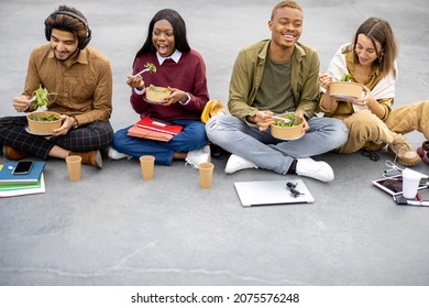 Multiracial Students Sitting And Eating Salad During Lunch On Asphalt At University Campus. Concept Of Education And Learning. Idea Of Student Lifestyle. Young Smiling Friends At Food Break