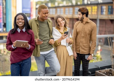 Multiracial students talking and using digital devices on balcony at university campus at day. Concept of education. Remote and e-learning. Idea of student lifestyle and friendship