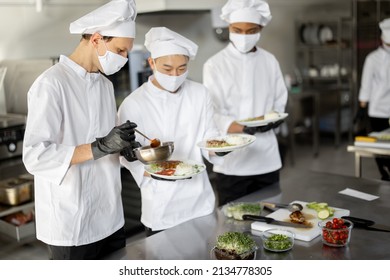 Multiracial Team Of Cooks Standing With Ready Meals For A Restaurant In The Kitchen. Chefs Wearing Uniform And Face Mask During Pandemic. Asian Chef With Latin And European Guys