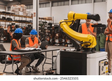 Multiracial team of industrial engineers programming a robotic arm in a large modern factory, discussing and analyzing data on a laptop while another engineer supervises the operation