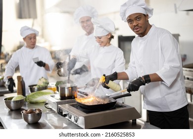 Multiracial team of professional cooks in uniform preparing meals for a restaurant in the kitchen. Latin guy burning pan, european cooks making sauce and asian chef managing the process. Teamwork and