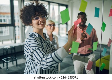 Multiracial Team At Work Writing Strategy Ideas On Sticky Notes On The Glass Wall While Having A Meeting In The Office. Business Project Planning. Focus On A Woman With Afro Hair.