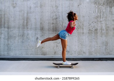 Multiracial teenage girl riding a skateboard in front of concrete wall, standing in one leg, balancing. Side view. - Powered by Shutterstock