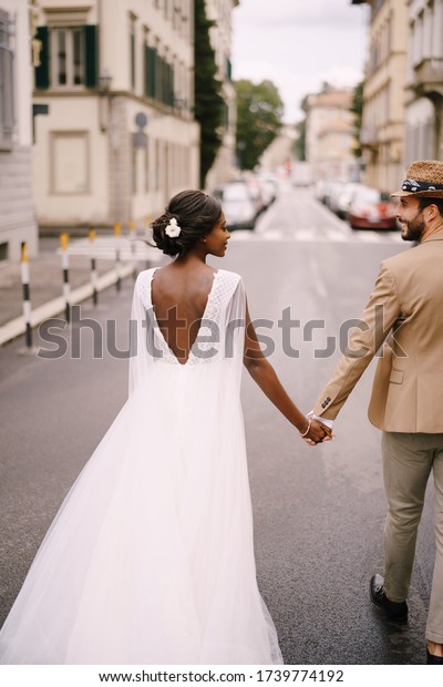Multiracial wedding couple. Wedding in\
Florence, Italy. African-American bride in a white dress and\
Caucasian groom in a hat are walking along the road among\
cars.