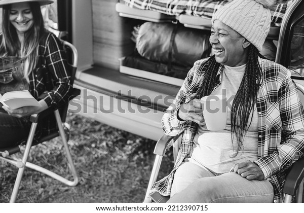 Multiracial women friends having fun\
camping with camper van while reading and drink coffee outdoor -\
Focus on african female face - Black and white\
editing.
