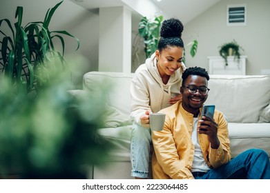 Multiracial young couple sitting on a comfortable sofa using smartphone and holding cups of hot beverages. Spouse relaxing together on cozy couch and online shopping on a mobile phone. Copy space. - Powered by Shutterstock