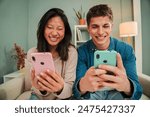 Multiracial young couple using their cellphones sitting on a couch at home living room. Boyfriend and girlfriend sharing and messaging on the social media. Two friends having fun with smart phone app