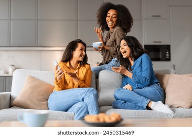 Multiracial young girlfriends share a laugh over coffee in a homey living room, enjoying casual camaraderie - Powered by Shutterstock