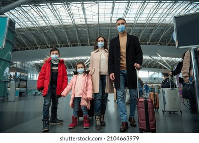 Multiracial Young Happy Family At The Airport With Luggage While Waiting For A Flight In A Pandemic. Winter Air Travel Concept.