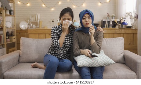 Multiracial Young People Watching Sad Movie In TV Relax On Sofa With Home Kitchen In Background In Bright Cozy Apartment. Two Asian Chinese And Muslim Women Friends Upset Crying Using Napkin Tissue