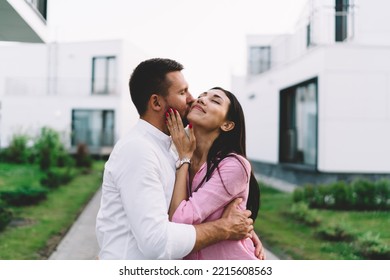 Multiracial Young Romantic Couple In Love Standing On Pathway While Boyfriend Holding Eyes Closed Girlfriend With Both Hands And Kissing On Cheek Outside