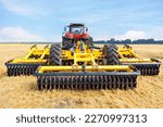 A multi-row disc harrow is hitched to an agricultural tractor against the background of a summer harvested wheat field and a blue sky.