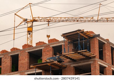 A multi-story brick building under construction with scaffolding-like wooden platforms and a large yellow tower crane in the background against a cloudy sky.