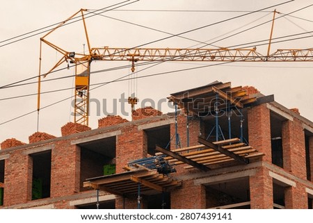 A multi-story brick building under construction with scaffolding-like wooden platforms and a large yellow tower crane in the background against a cloudy sky.