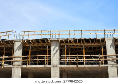 Multi-story building under construction, featuring concrete pillars, wooden formwork for floor slabs, and temporary yellow support props beneath a clear blue sky with copyspace