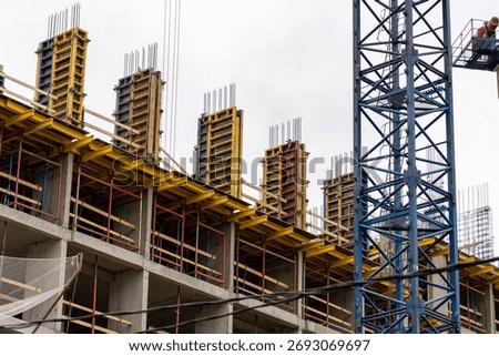 Multi-story building under construction with yellow formwork, exposed rebar, and prominent blue tower crane against overcast sky, depicting urban development progress
