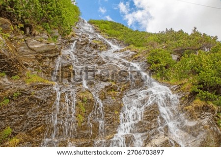 Multi-stream waterfall running over a broad rocky slope with alpine shrubs and cloudy sky.