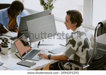 Multitask Caucasian programmer using gray laptop to search information while using computer to write code, his African American coworker writing report in blurred background
